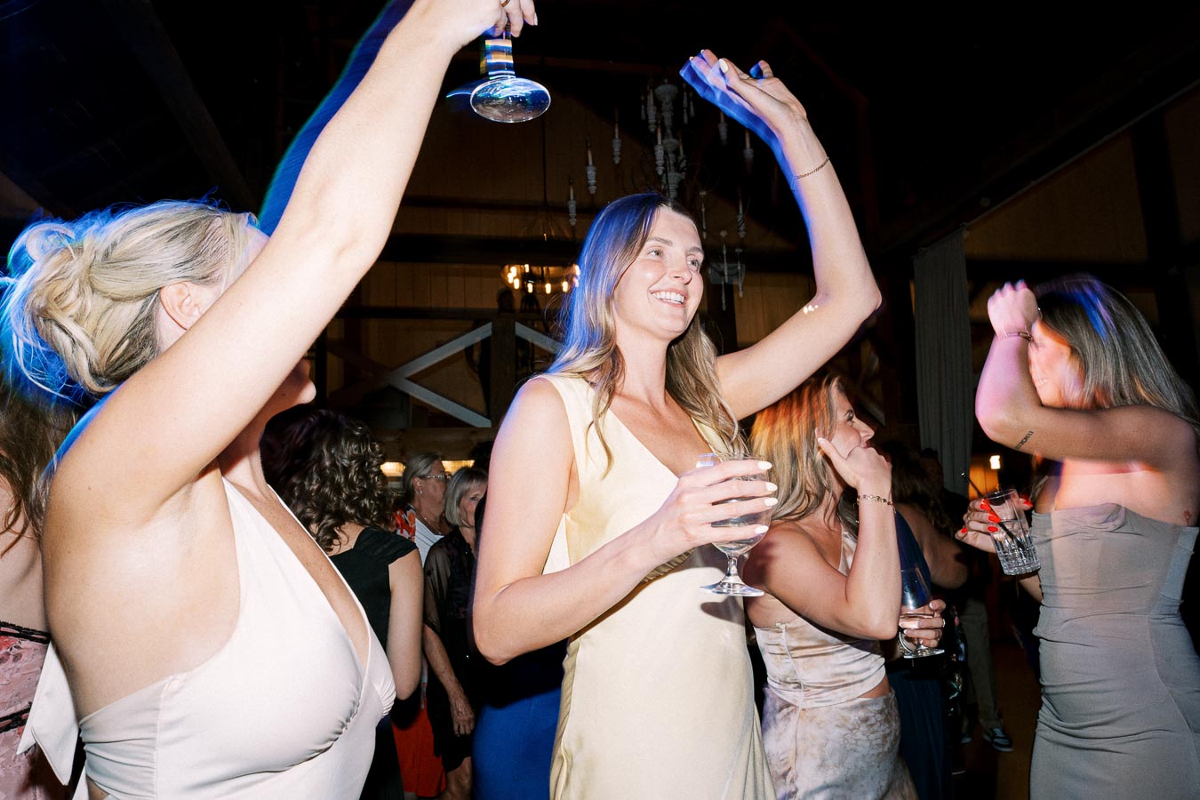 Women enjoying a lively party, dancing and smiling with drinks in hand under festive lighting.
