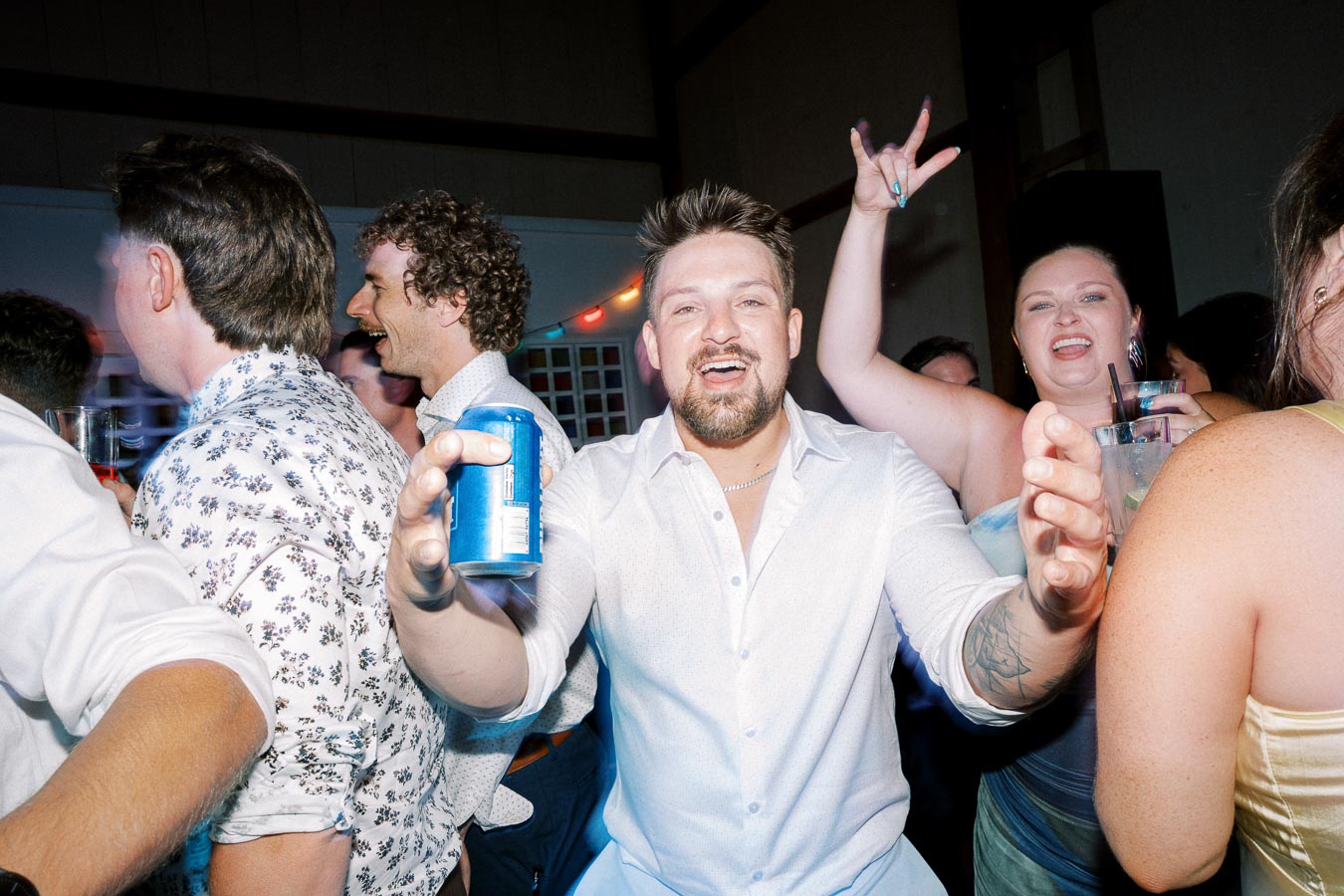 A group of people celebrating at a party, with a man in a white shirt holding a blue can, surrounded by other cheerful guests enjoying drinks and dancing under colorful lights.