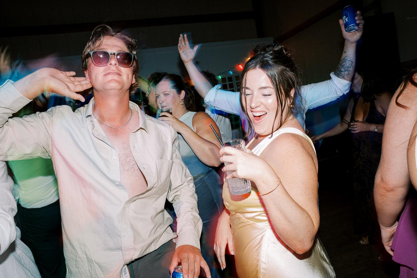 A group of people enjoying a lively party, with a man in sunglasses and an unbuttoned shirt dancing energetically alongside a woman in a gold dress holding a drink, surrounded by other partygoers in a colorful, festive atmosphere.