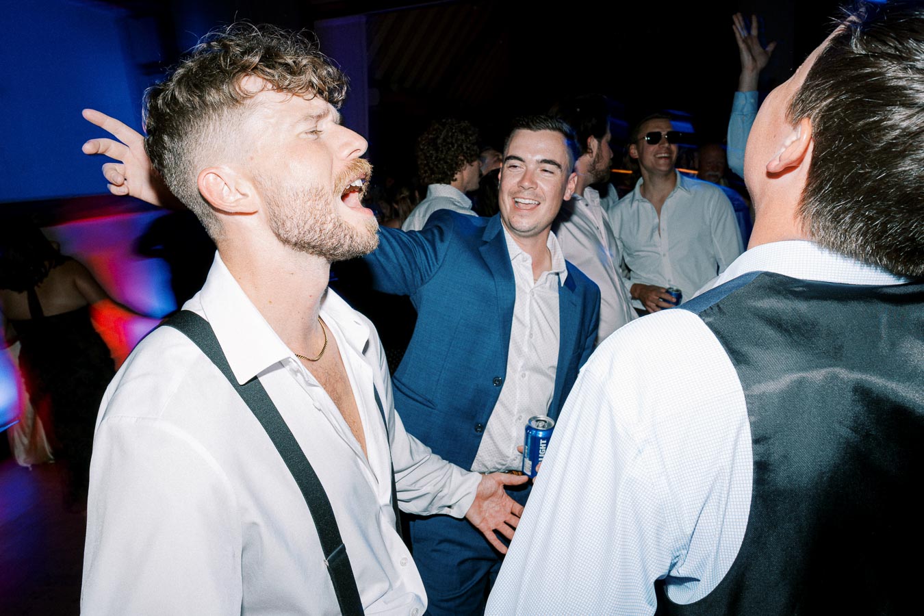 Men enjoying a lively party scene, dressed in formal shirts, with one man singing energetically in the foreground, creating a festive atmosphere.