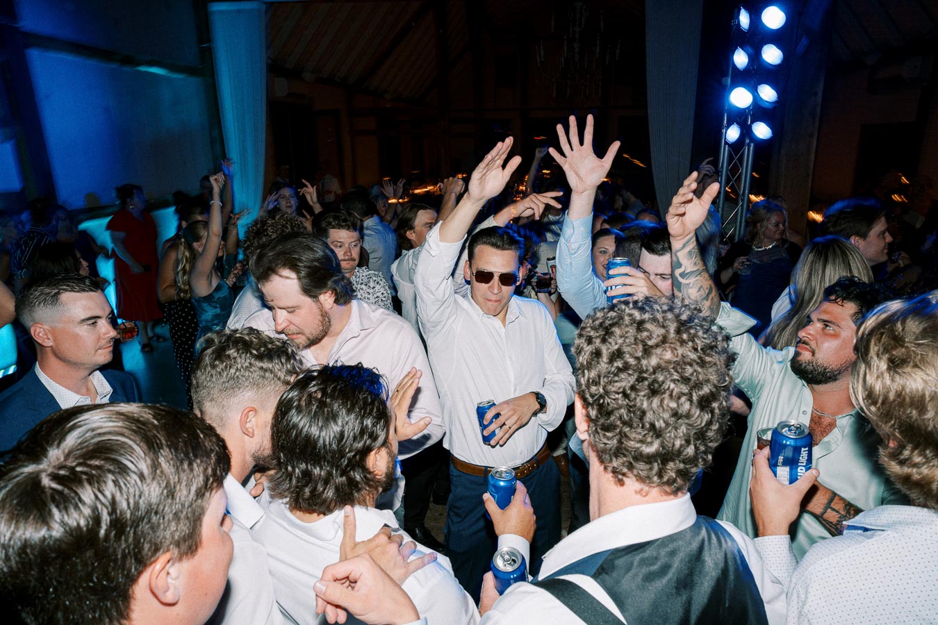 A lively group of people dancing at a party, with some holding drinks and others raising their hands in celebration, under vibrant blue lighting.