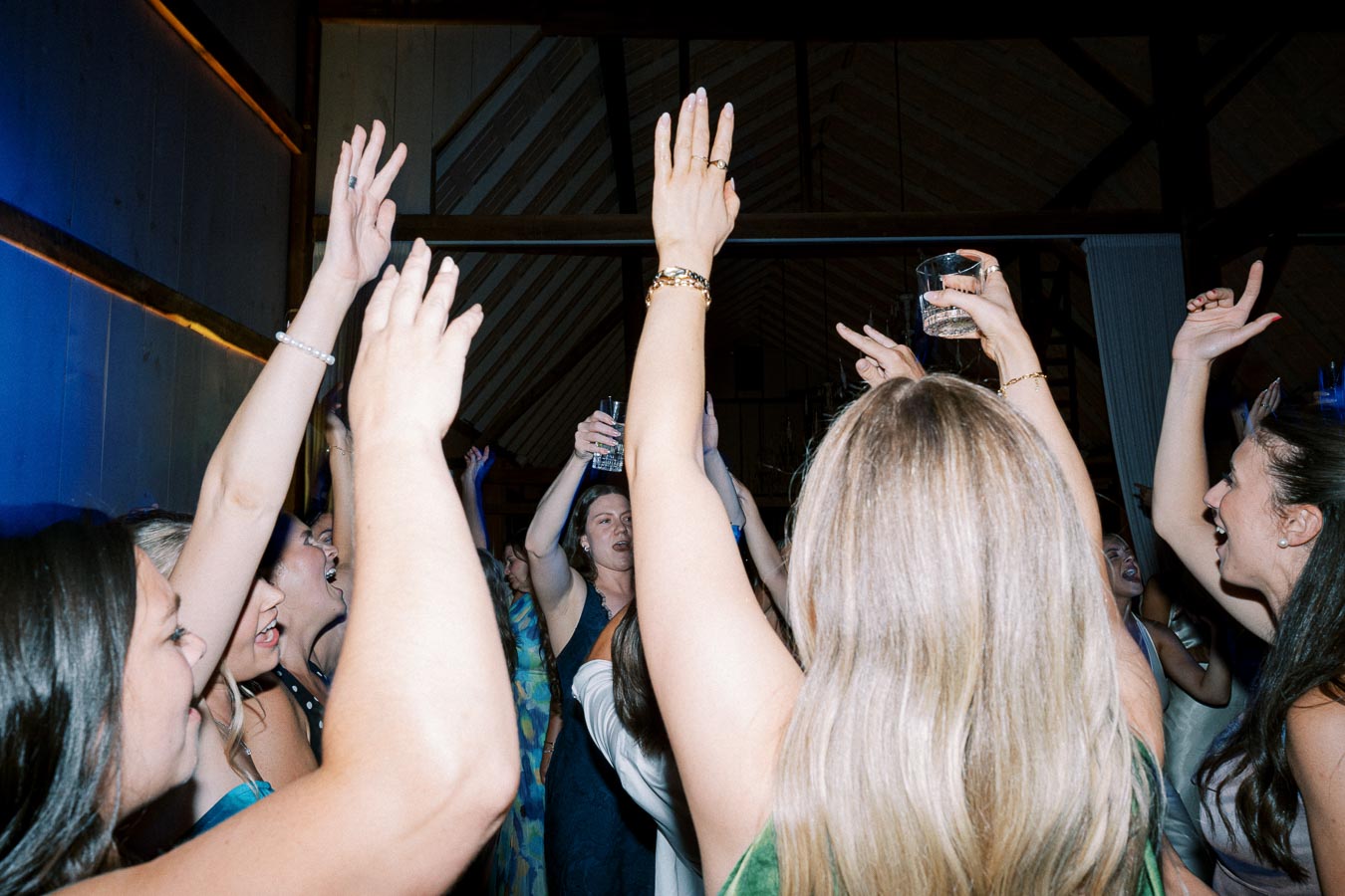 Group of people dancing and celebrating indoors with raised hands and drinks at a lively party.