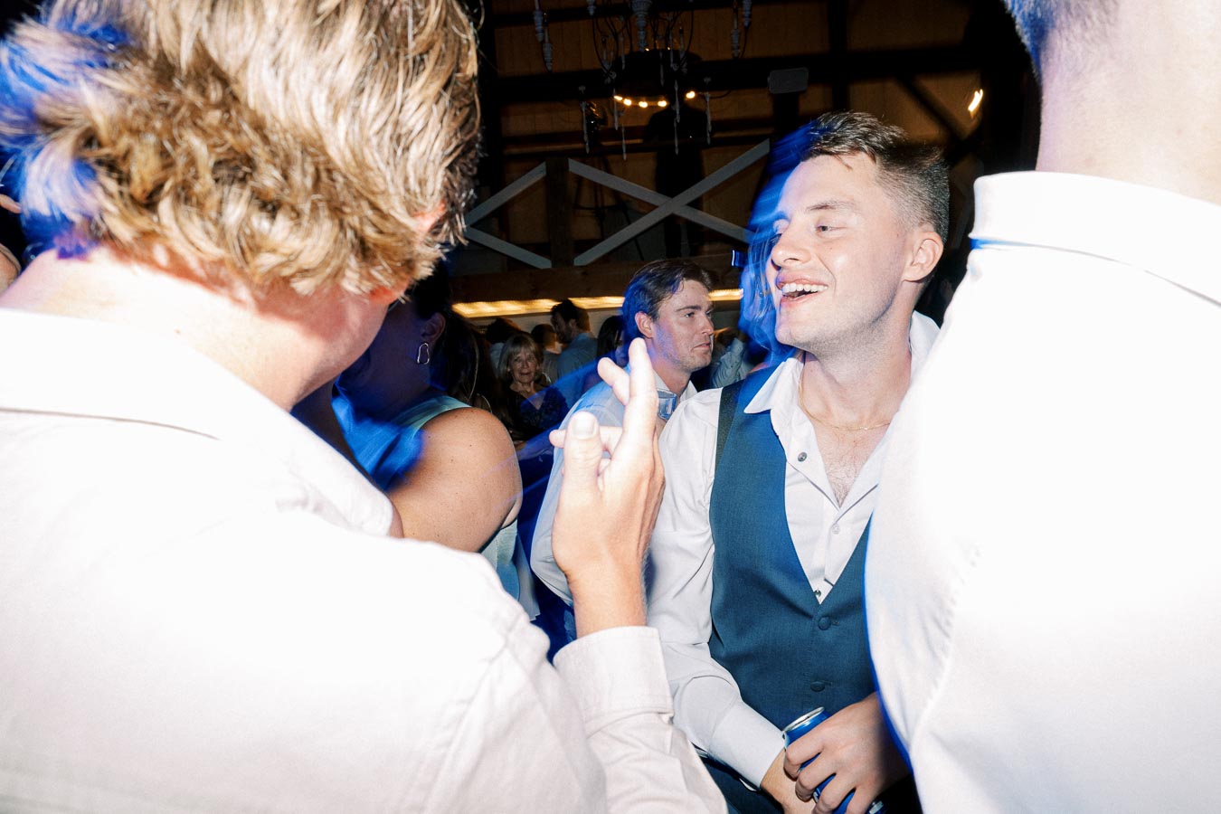 A group of people enjoying a lively party, with a focus on a smiling man in a white shirt and vest, surrounded by guests in a warmly lit indoor venue.