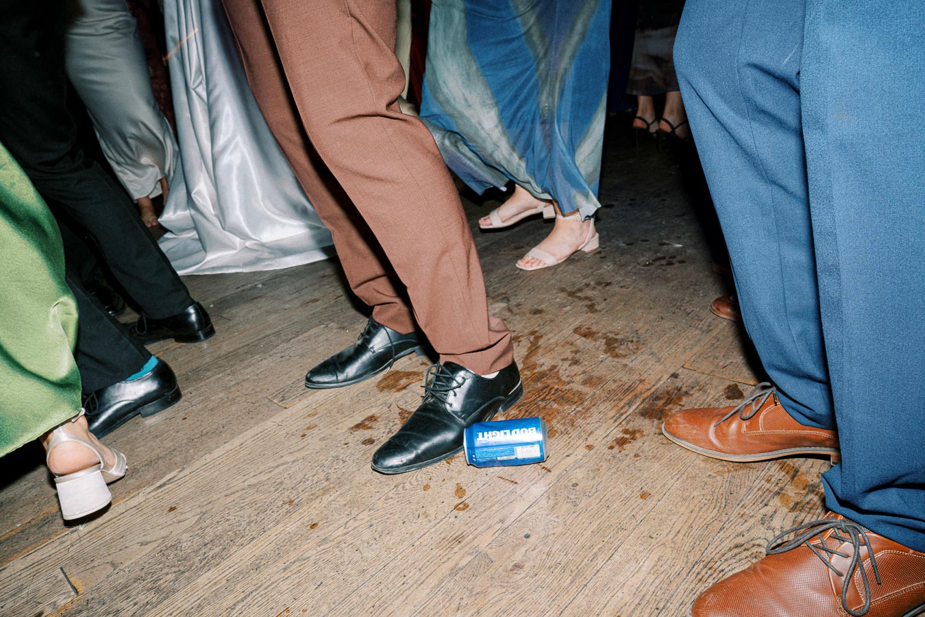 Close-up of people dancing in elegant attire on a wooden floor, with a discarded beverage can among them, capturing a lively party atmosphere.