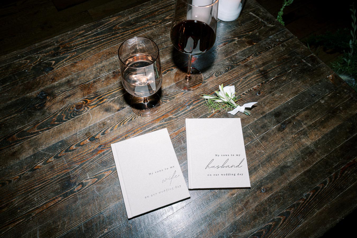 Wedding vow books on a rustic wooden table with wine and water glasses nearby, accompanied by a small sprig of greenery.