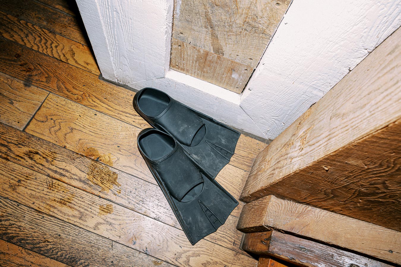 Black swim fins placed on wooden floorboards in a corner, next to white and wooden walls.