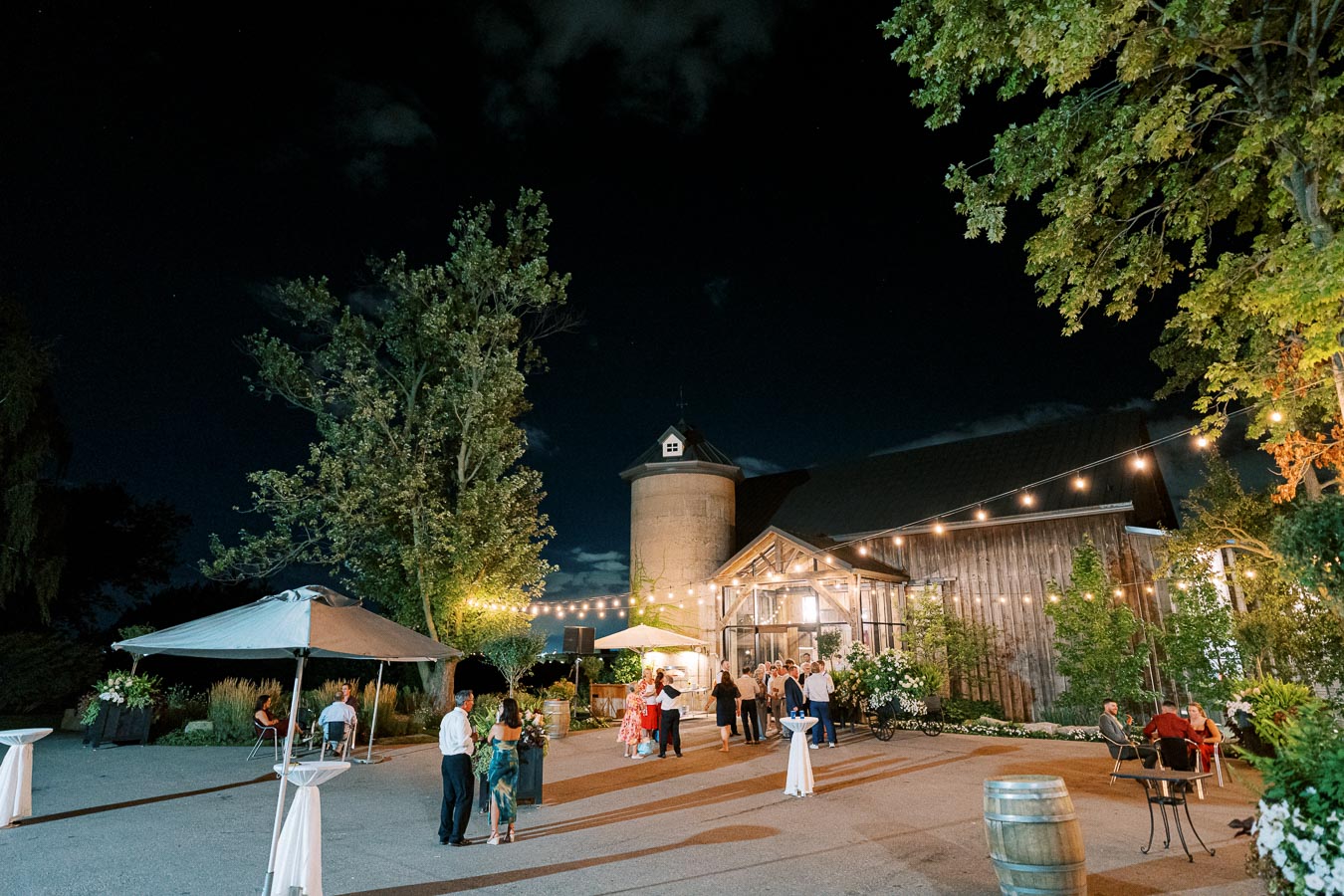 Nighttime outdoor wedding reception at a rustic barn venue, featuring string lights, guests mingling, and a tree-lined backdrop.