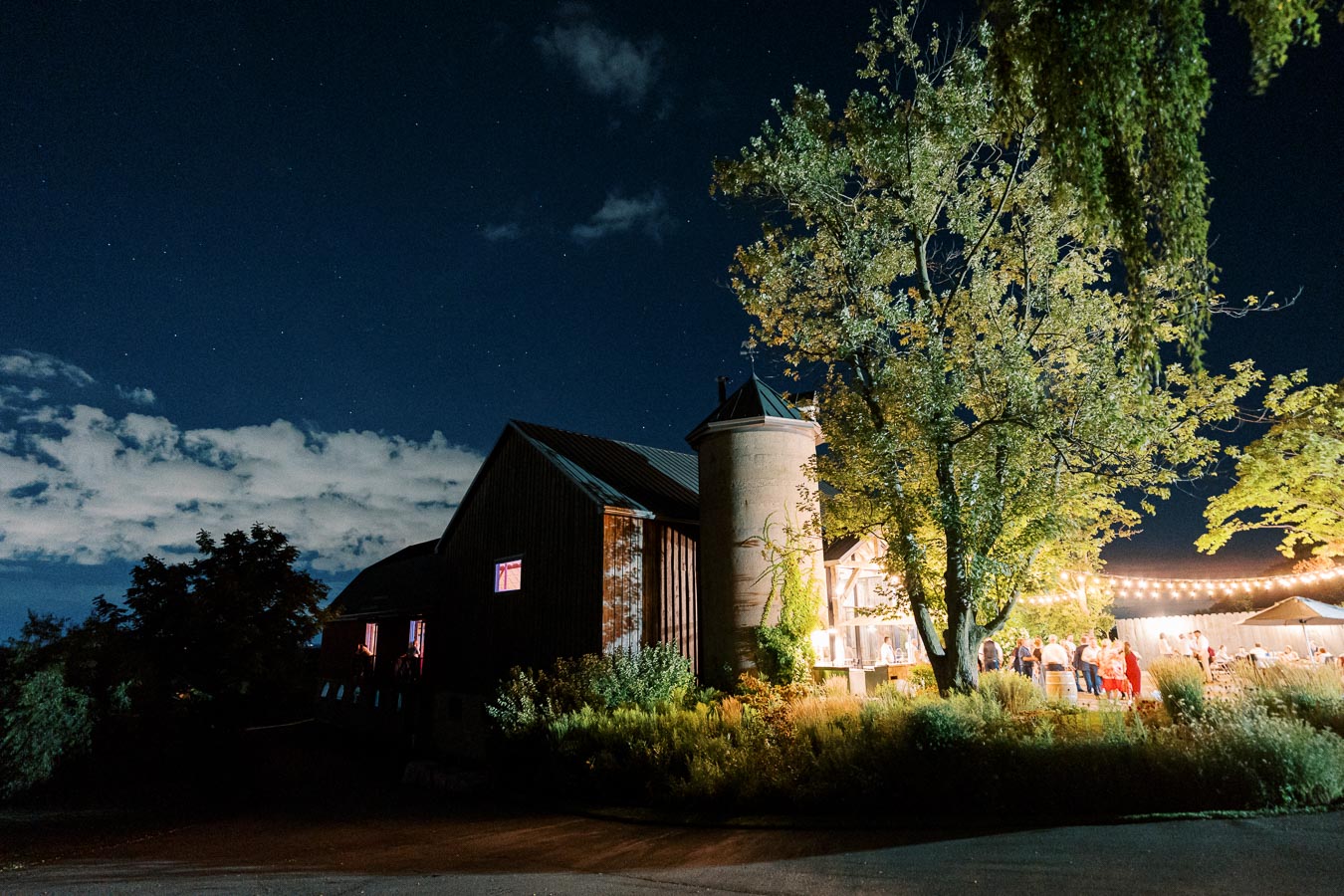 A beautifully illuminated rustic barn venue at night with a large tree in the foreground, string lights, and guests gathered outside under a starry sky.