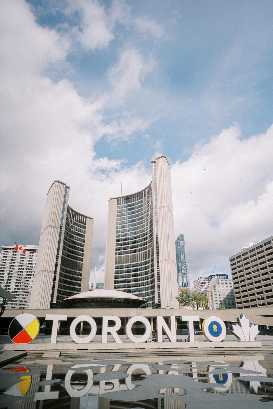 Skyline view of Toronto City Hall with large Toronto sign in foreground and cloudy blue sky, showcasing iconic architecture at Nathan Phillips Square.