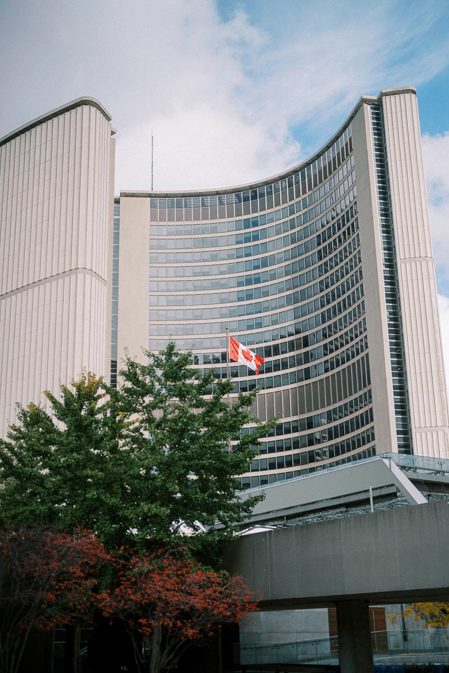 Modern glass-clad city hall building with Canadian flag and trees in the foreground under a partly cloudy sky.