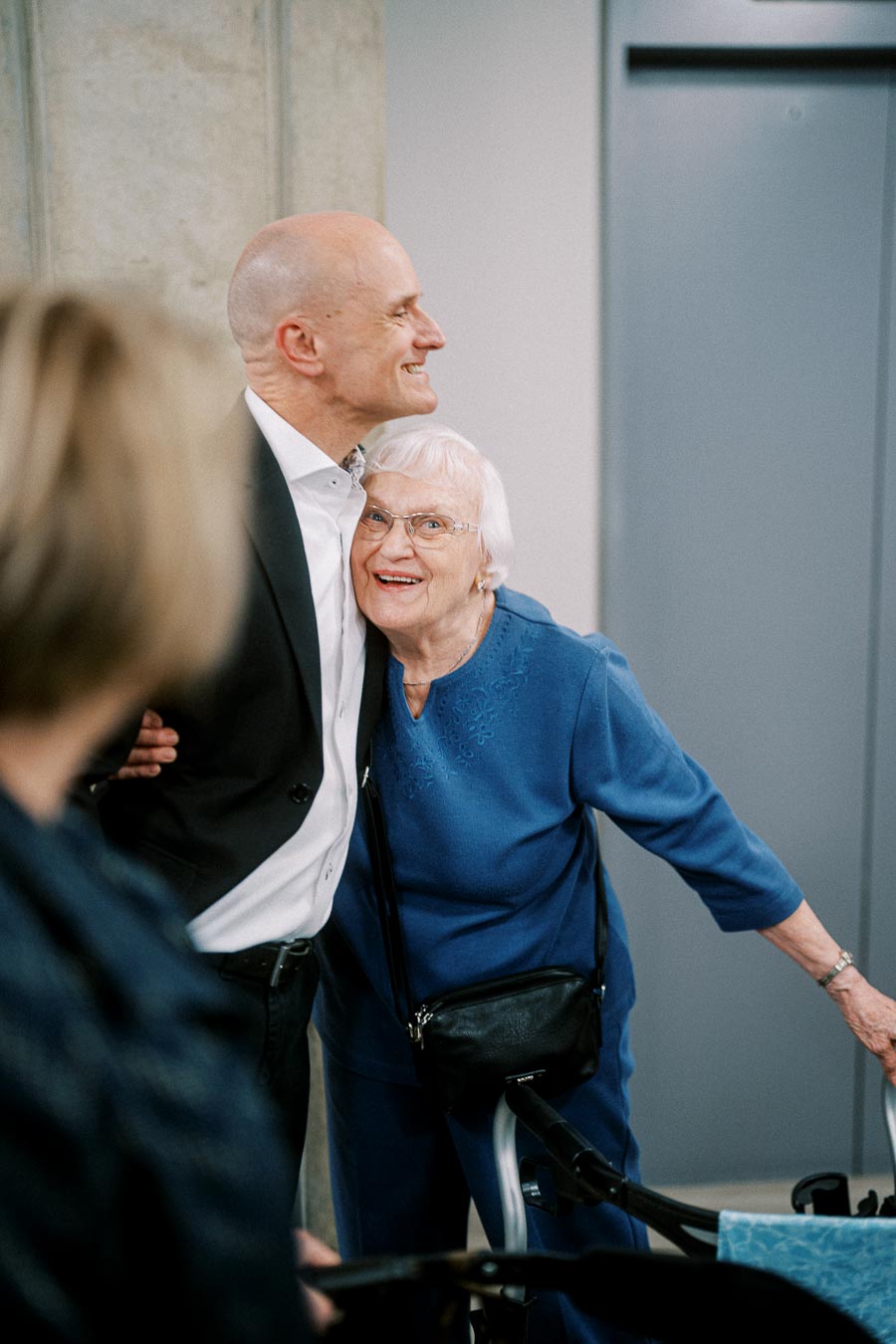 Joyful elderly woman in blue embraces man in a suit indoors, showcasing intergenerational bond and happiness.