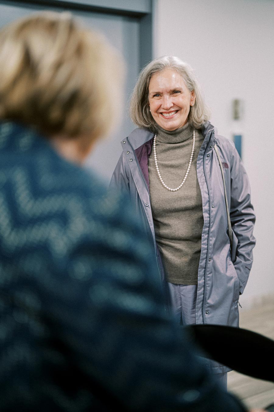 Smiling woman in a gray jacket and turtleneck, wearing a pearl necklace, engaged in conversation indoors.