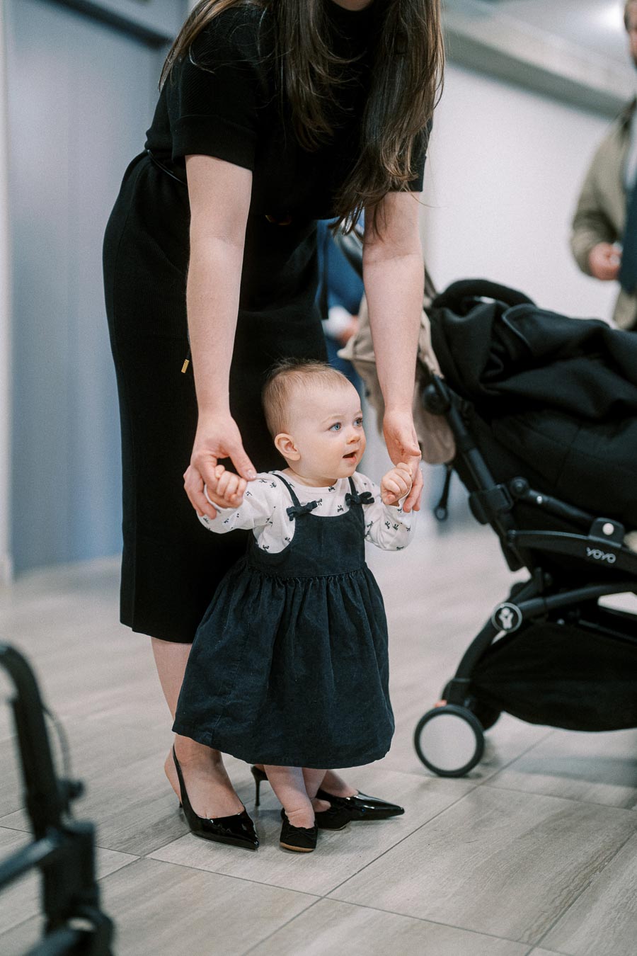 A young mother helps her baby take first steps indoors, next to a stroller, highlighting parent-child bonding and early childhood development.
