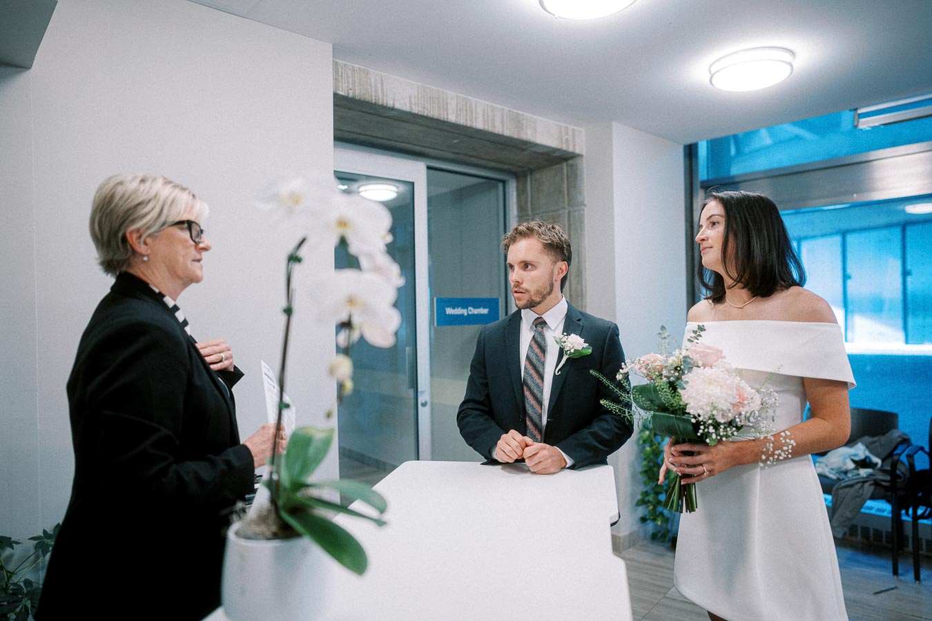 A couple in formal attire standing at a counter in a wedding registry office, speaking with an official. The groom is in a suit, and the bride is wearing a white dress holding a bouquet of flowers, with a sign reading Wedding Chamber visible in the background and a white orchid plant in the foreground.