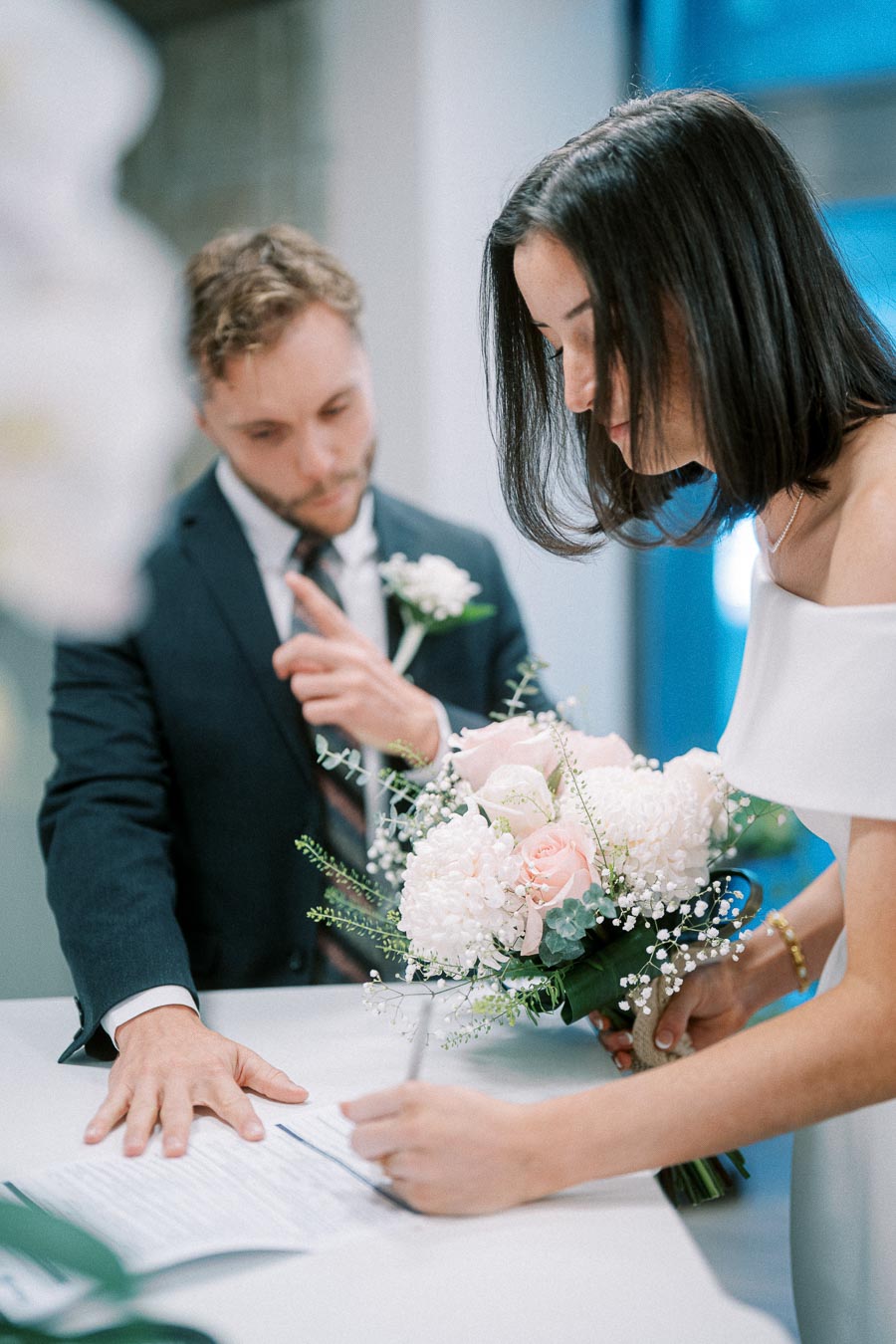 A couple signs a wedding certificate on a table, with the bride holding a bouquet of pink and white flowers, and the groom assisting, both dressed in formal attire.