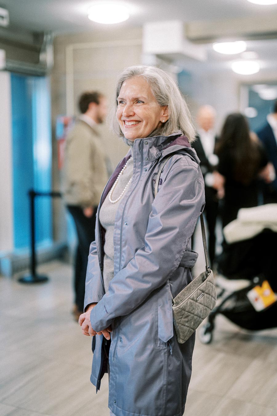 Elderly woman smiling in a public indoor setting, wearing a purple jacket and a pearl necklace, with a quilted handbag.