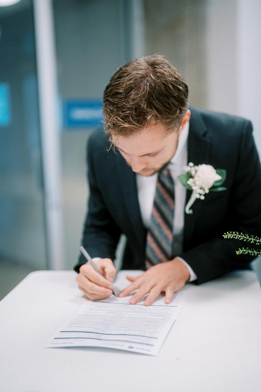 Man in a suit signing a document at a table, wearing a striped tie and a boutonniere, in a professional setting.