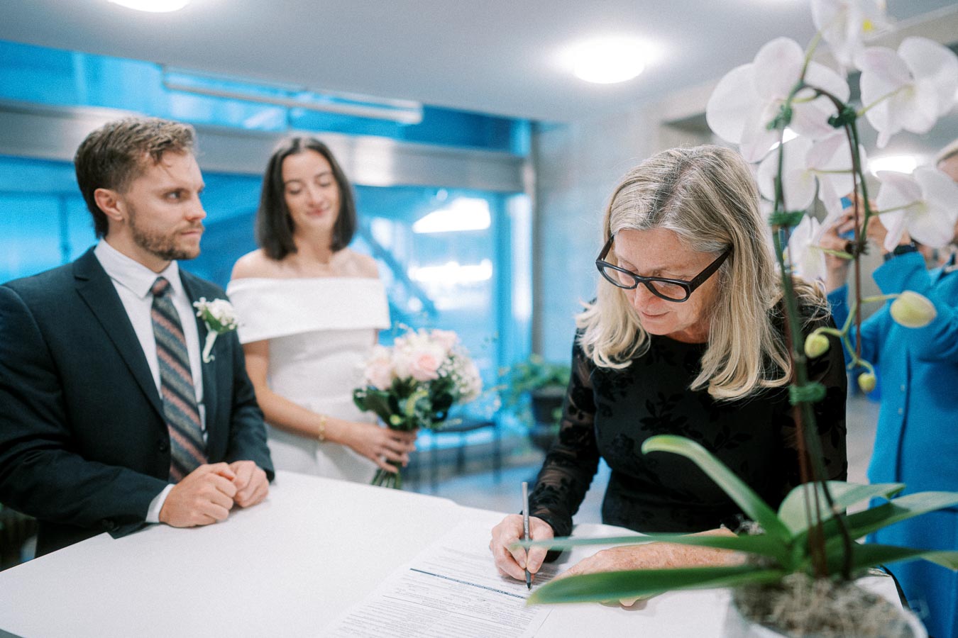 A couple in wedding attire standing at a table while a woman in glasses signs a document, with flowers in the foreground, capturing a candid moment of a civil marriage ceremony indoors.