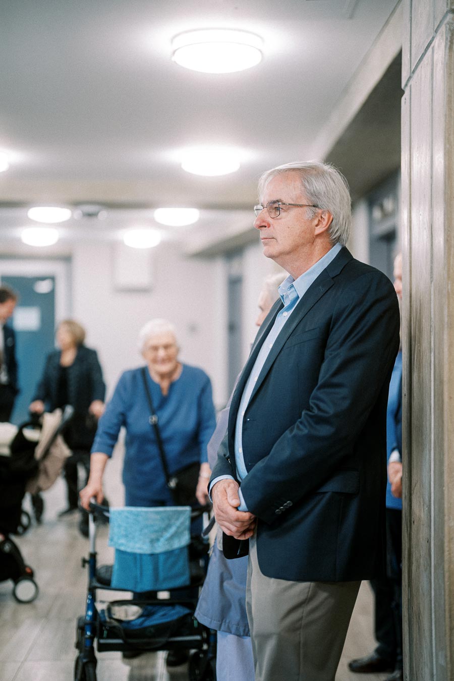 Elderly individuals gathered in a well-lit hallway, with one man standing against a wall while others with walkers converse in the background, depicting a community or social gathering scene.