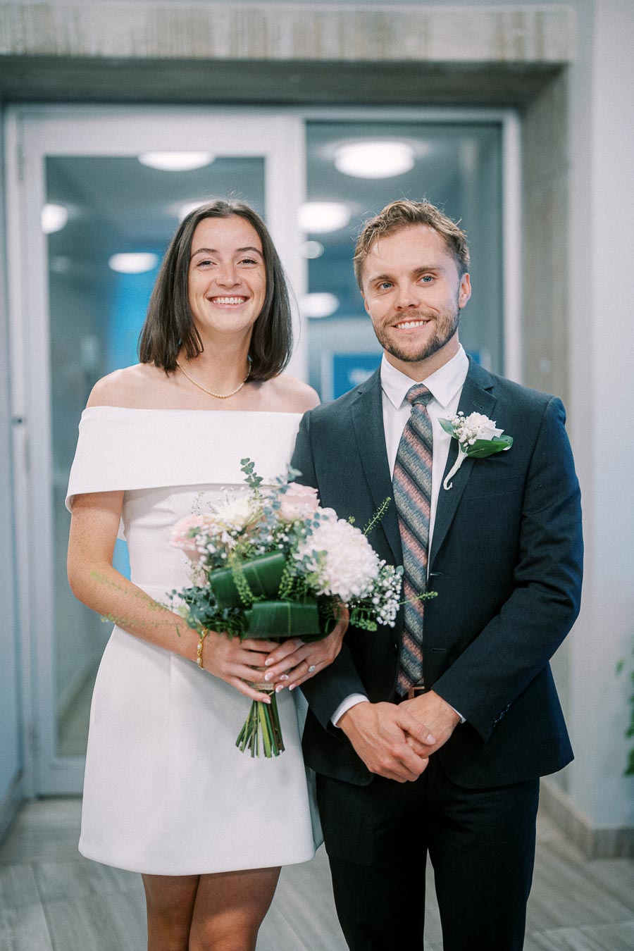 A smiling couple, elegantly dressed, with the bride in a white off-shoulder dress holding a bouquet of flowers, and the groom in a dark suit with a boutonniere, standing indoors in front of a modern building entrance.