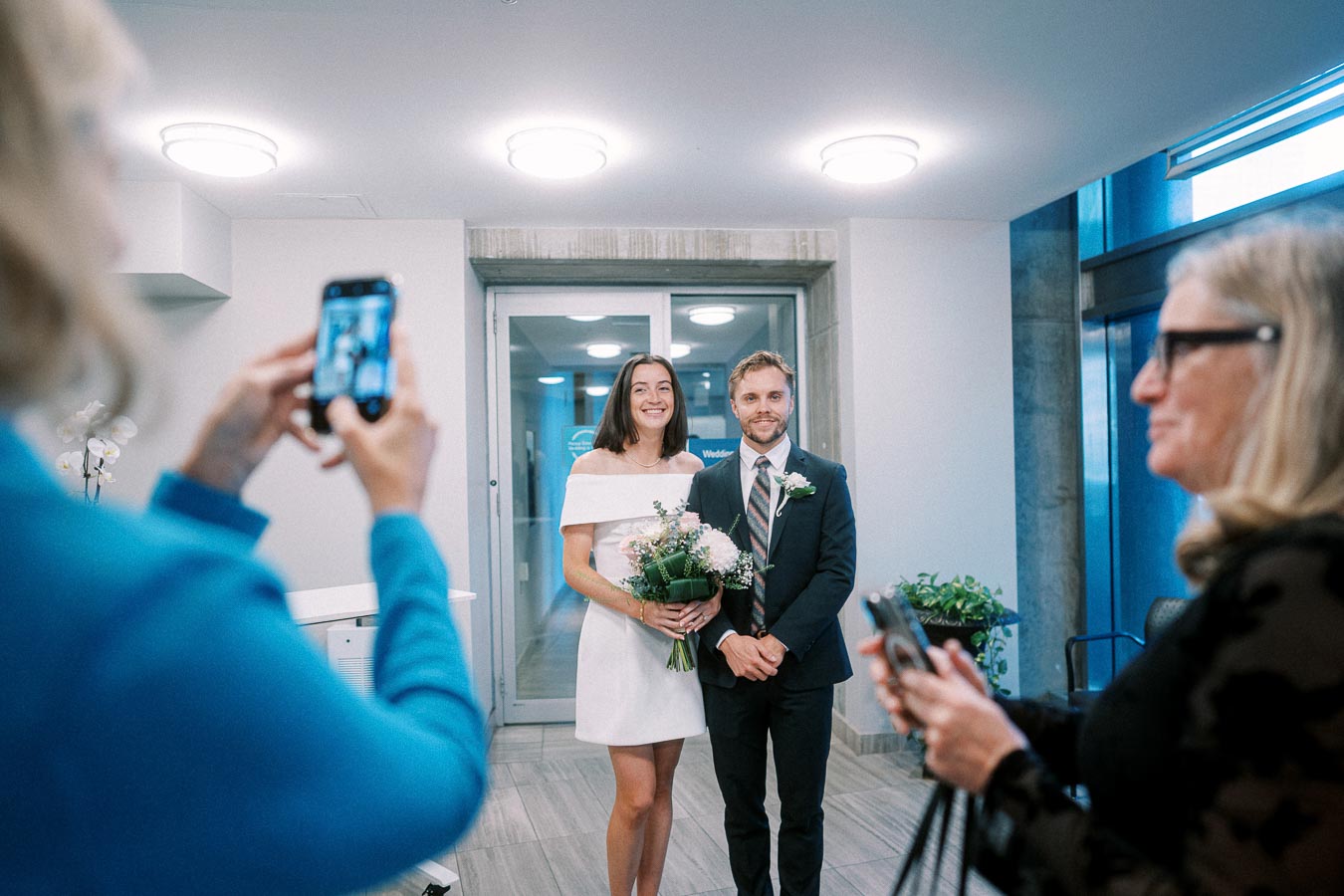 A bride and groom pose for photos in a modern, well-lit room. The bride holds a bouquet of flowers, wearing an elegant white dress, while the groom is in a dark suit with a floral boutonnière. Friends capture the moment on their smartphones, adding to the joyous atmosphere of the occasion.