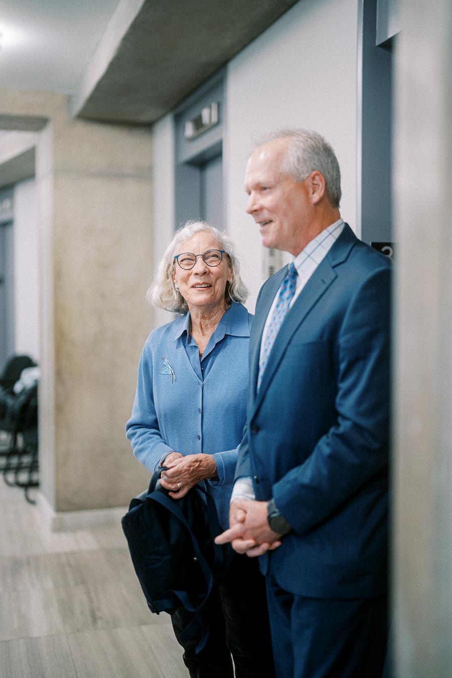 Elderly woman and man in formal attire smiling and conversing in a modern office hallway, showcasing a professional and friendly atmosphere.