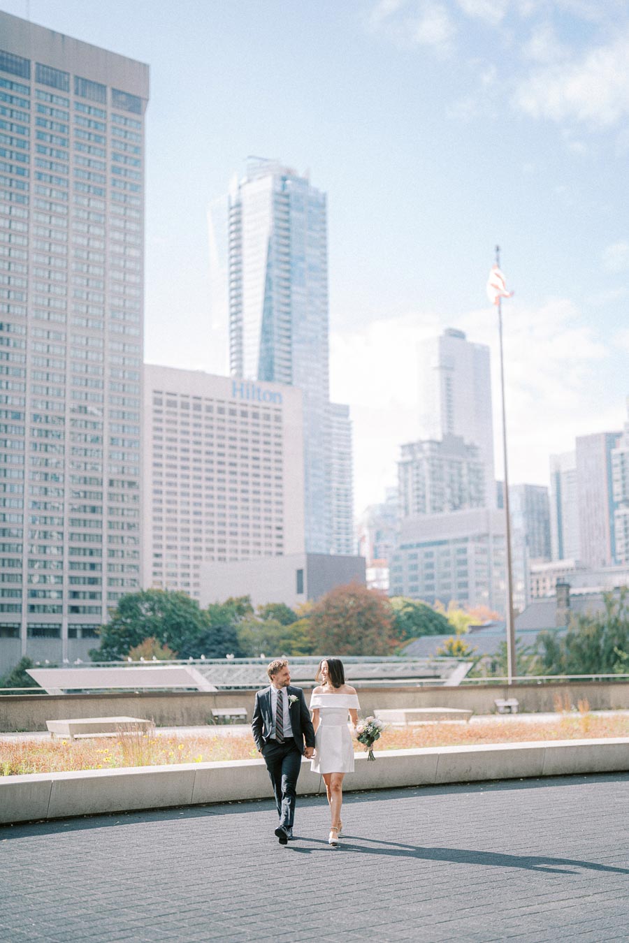 Couple walking hand in hand near skyscrapers in urban cityscape, man in suit and woman in white dress holding a bouquet, modern skyline and clear sky.