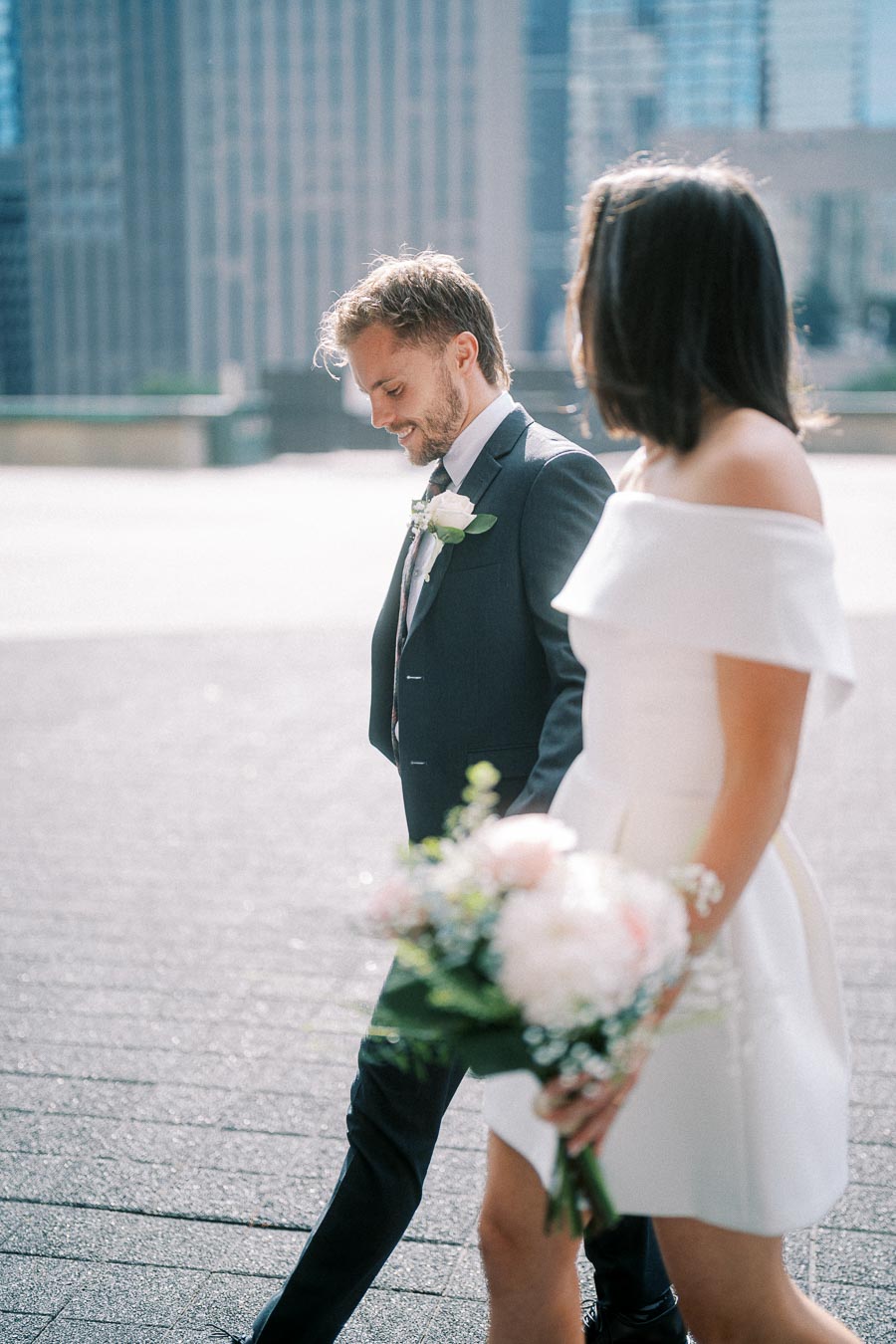A bride and groom walking outdoors in a city setting, with the bride holding a bouquet of white and pink flowers, and both wearing formal attire.
