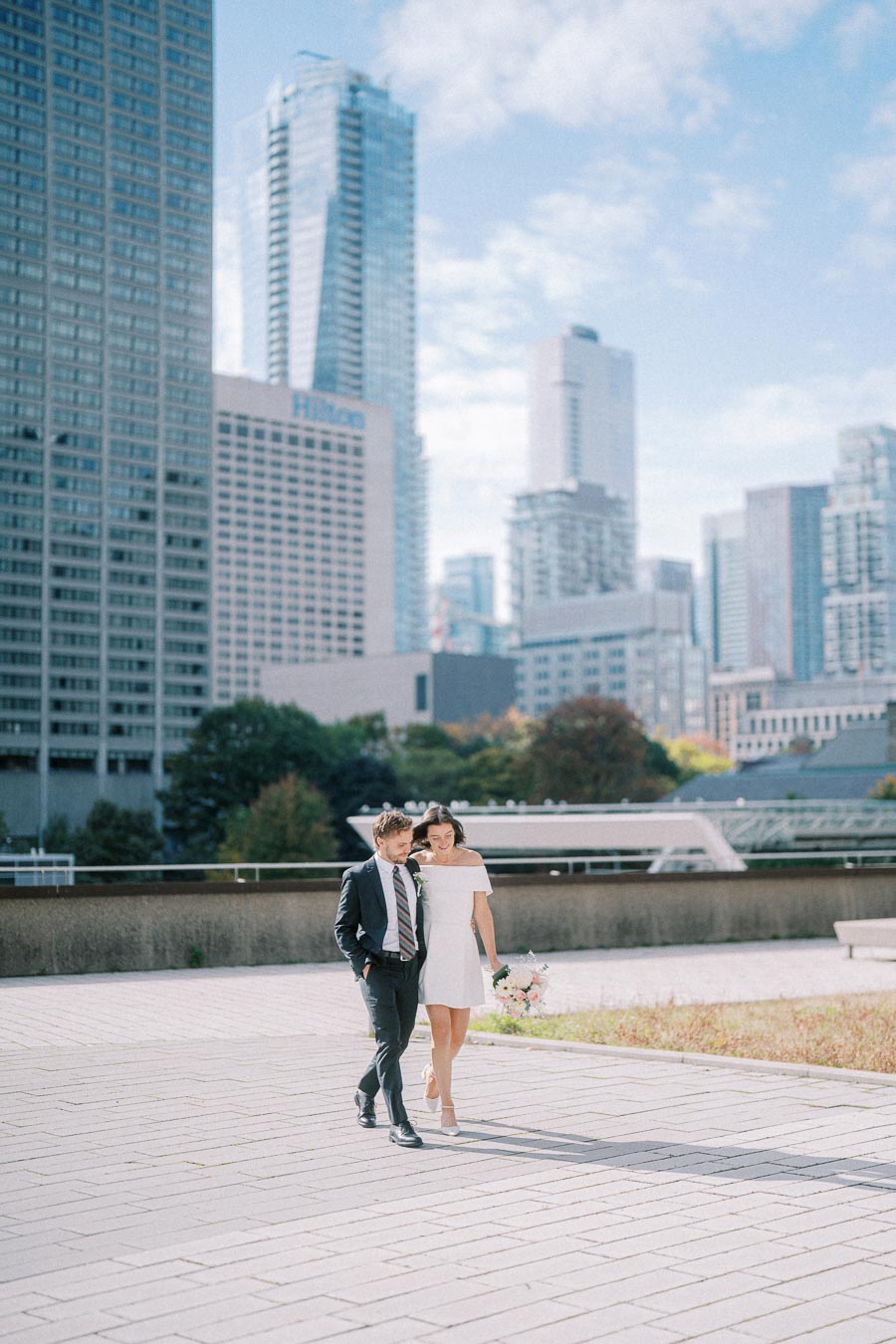Couple walking in cityscape, dressed for a wedding, surrounded by modern skyscrapers and clear blue sky