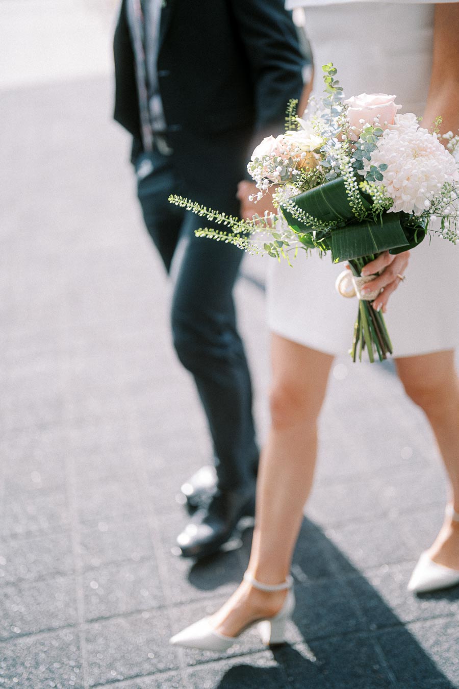 A bride holding a vibrant bouquet of white and pink flowers, walking with a man in a suit on a textured pavement.