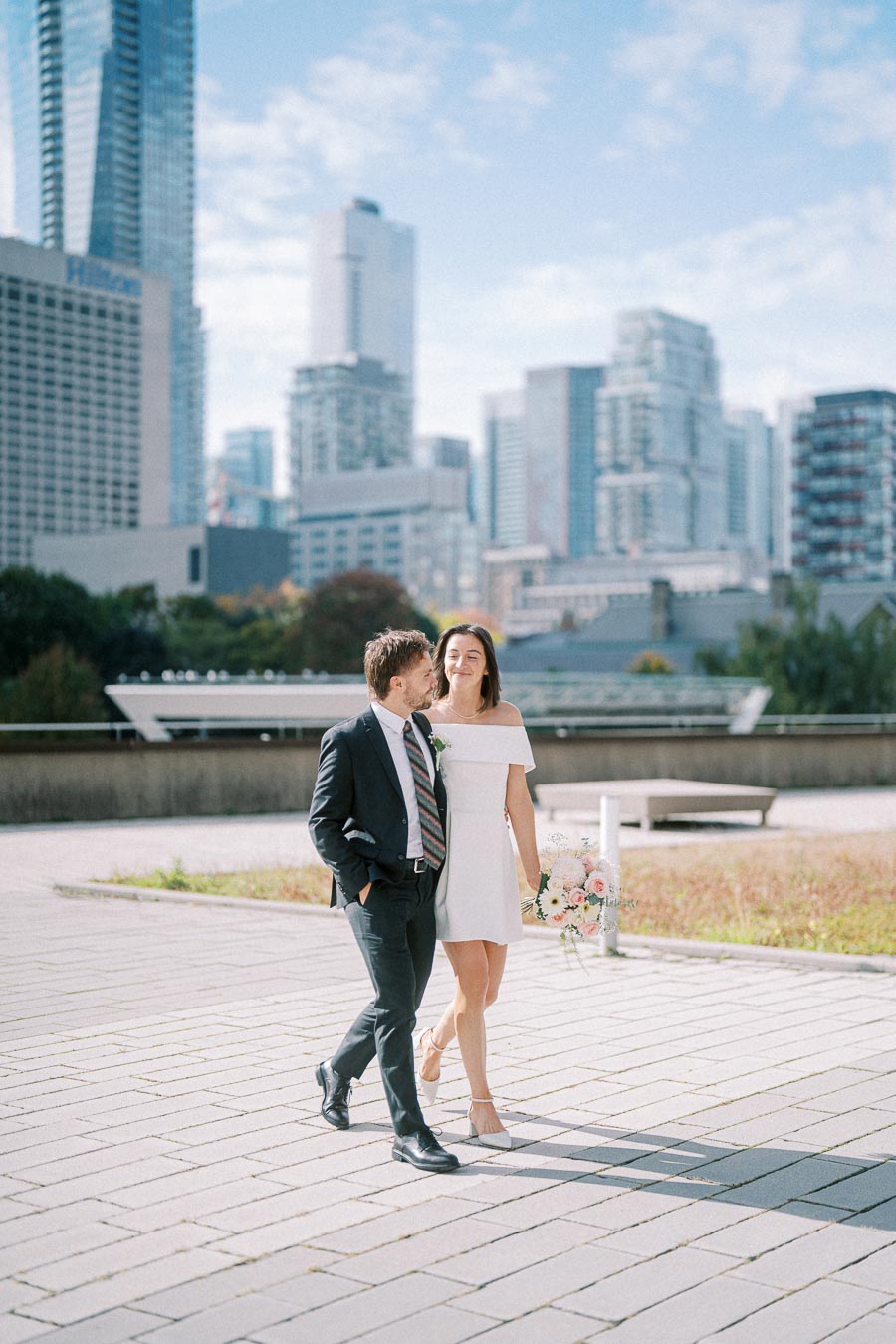 A couple in formal attire, with the woman holding a bouquet of flowers, walking along a paved pathway in an urban cityscape with skyscrapers in the background.