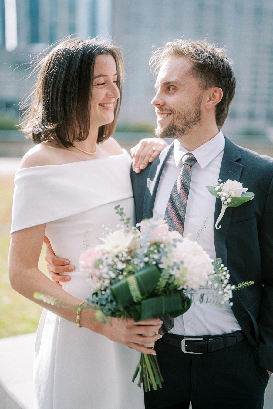 A bride in an elegant white dress holding a bouquet of flowers stands beside a groom in a dark suit with a boutonniere, both smiling at each other outdoors.