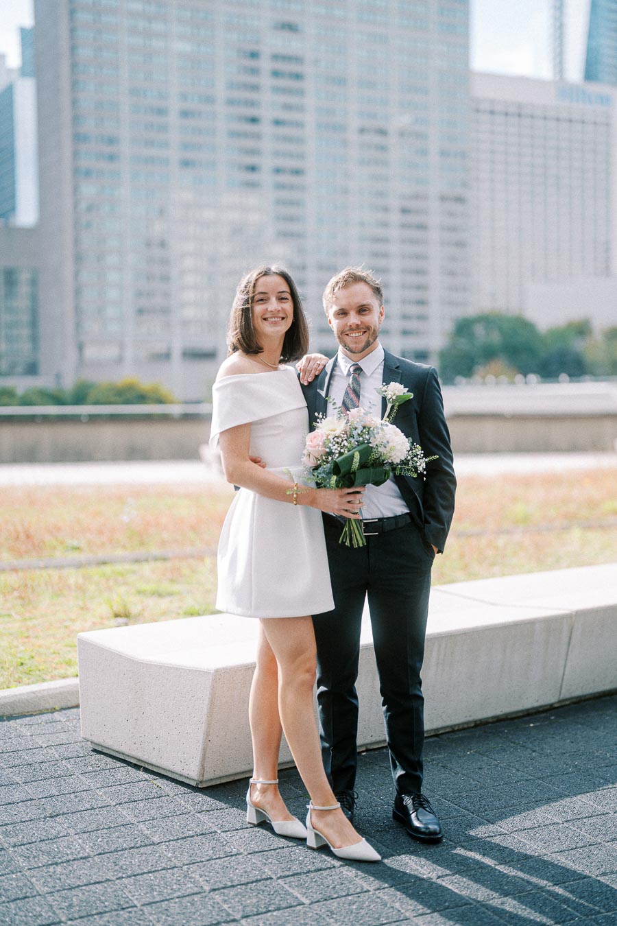 A smiling couple in formal attire, posing outside with a modern cityscape background; the woman holds a bouquet of flowers, wearing a white dress and heels, while the man is dressed in a dark suit and tie.