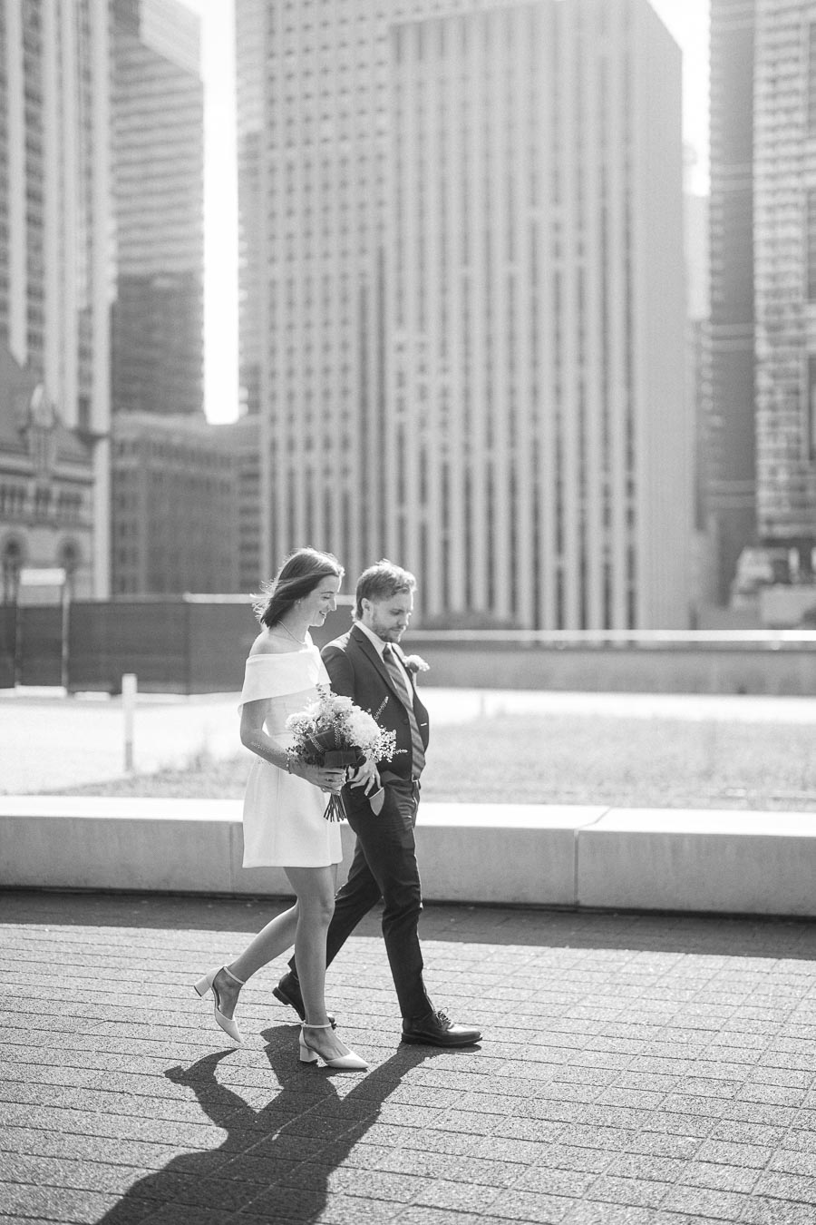 Black and white photo of a couple walking on a city street, with the woman holding a bouquet of flowers and tall buildings in the background.