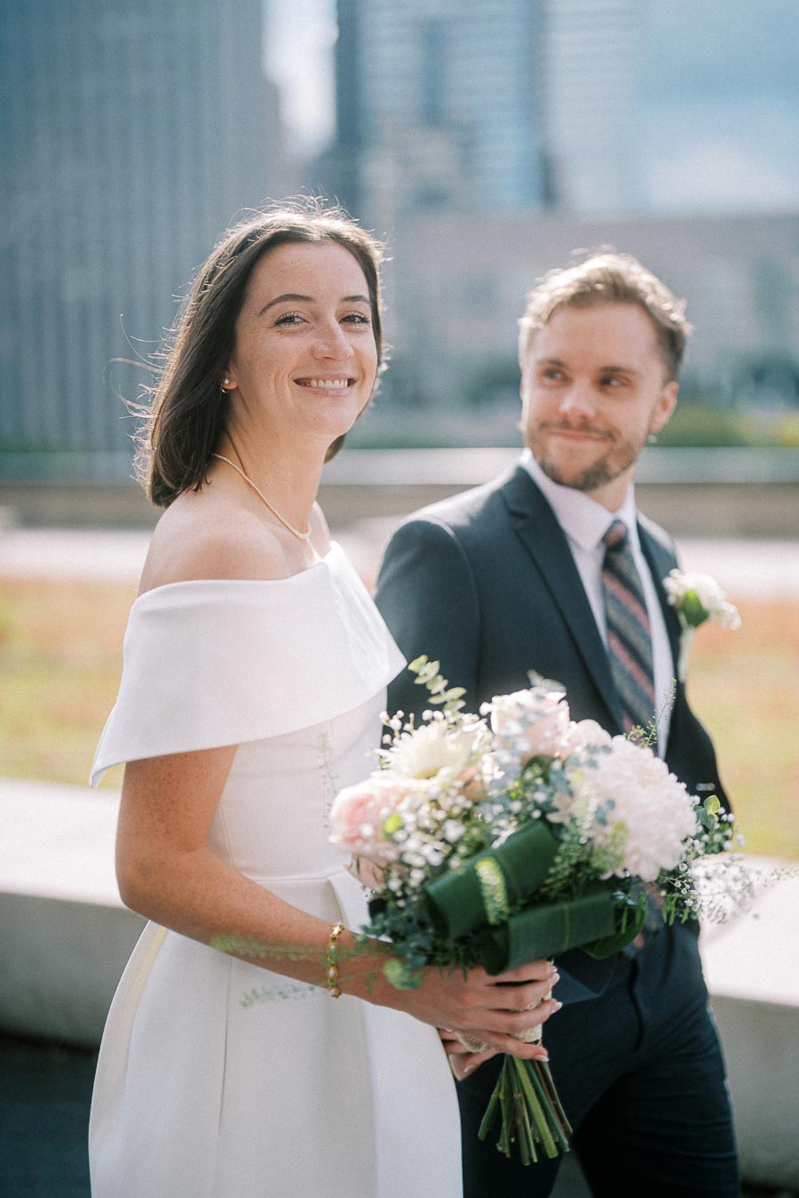Happy couple on their wedding day, bride in an elegant white dress holding a bouquet of flowers, standing beside groom in a dark suit with a cityscape background.