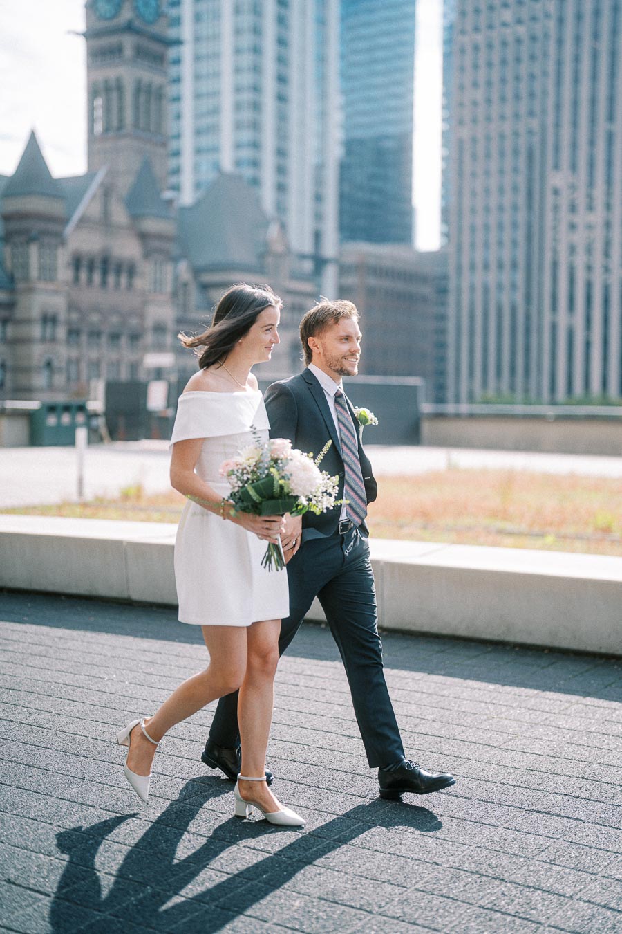 A couple walks side by side in formal attire, with a woman in a white dress holding a bouquet of flowers and a man in a suit, against a backdrop of historic and modern buildings in a city setting.