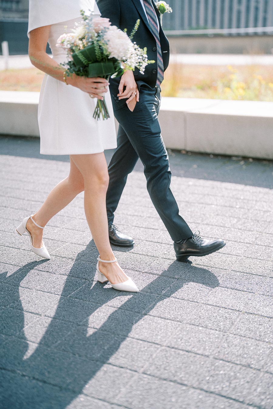 A bride in a white dress and heels walks beside a groom in a suit, holding a bouquet of flowers, casting shadows on a sunlit pathway.