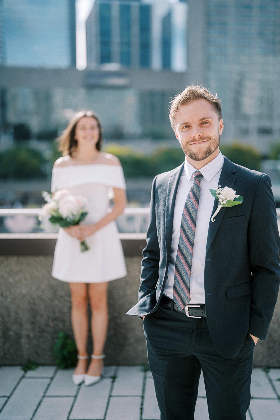 A groom in a suit is smiling confidently on a rooftop, with a bride in a white dress holding a bouquet standing in the blurred background, against a cityscape backdrop.