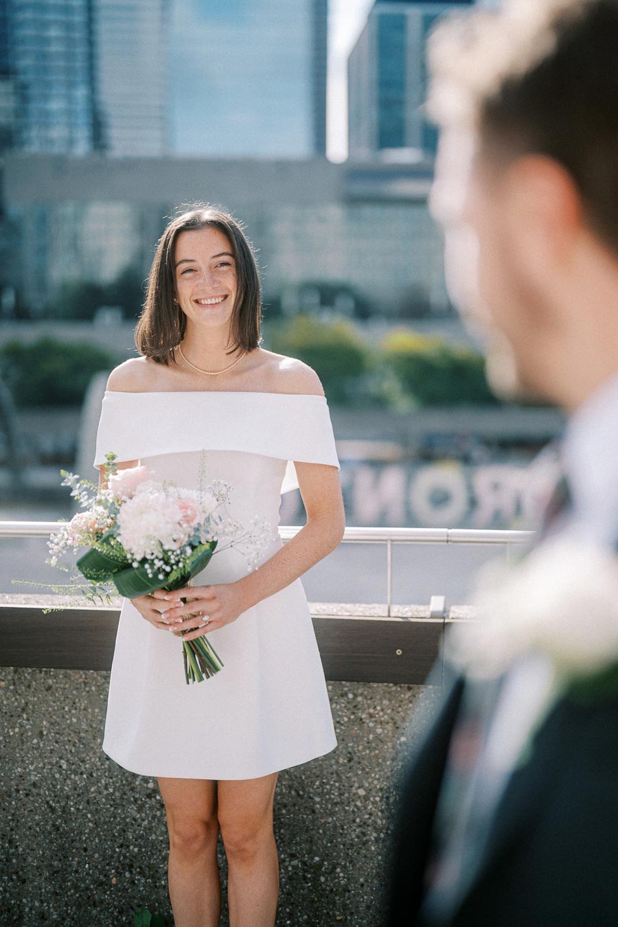 A bride in an off-the-shoulder white dress and holding a bouquet stands on a balcony with a cityscape in the background, smiling at a groom in a suit.