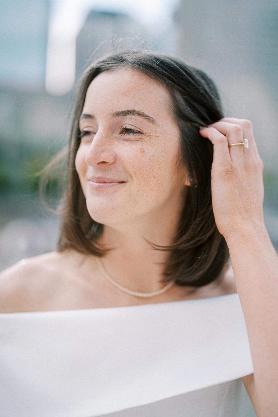 Smiling woman with brown hair wearing an off-shoulder white dress and necklace, adjusting hair with hand, outdoors with blurred cityscape background.