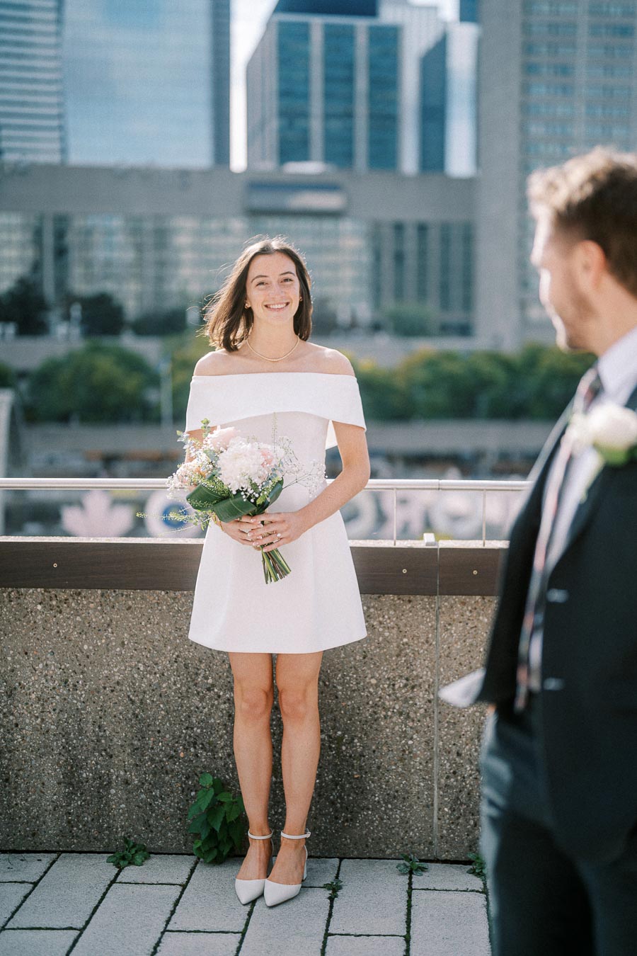 A smiling woman in a white off-shoulder dress holding a bouquet stands on a rooftop with a city skyline in the background, while a man in a suit looks on.