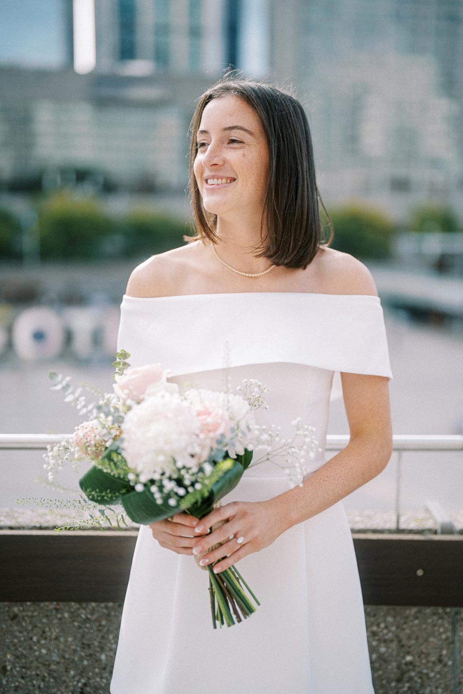 A smiling bride in an off-shoulder white wedding dress holds a bouquet of white and pink flowers, with a cityscape in the background.