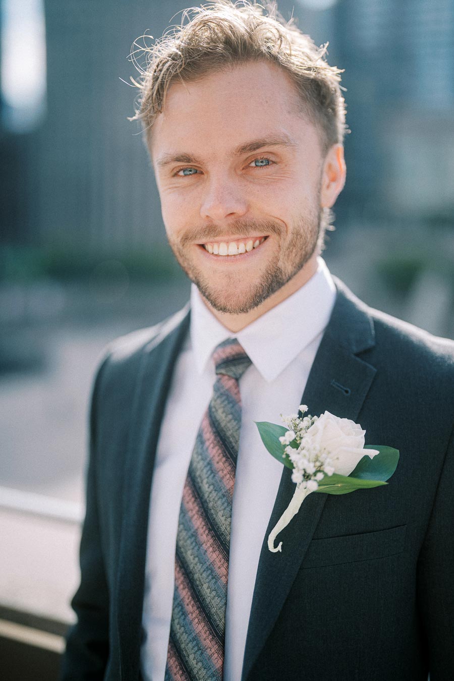 Smiling man in formal suit with floral boutonniere, standing outdoors with a blurred cityscape background.