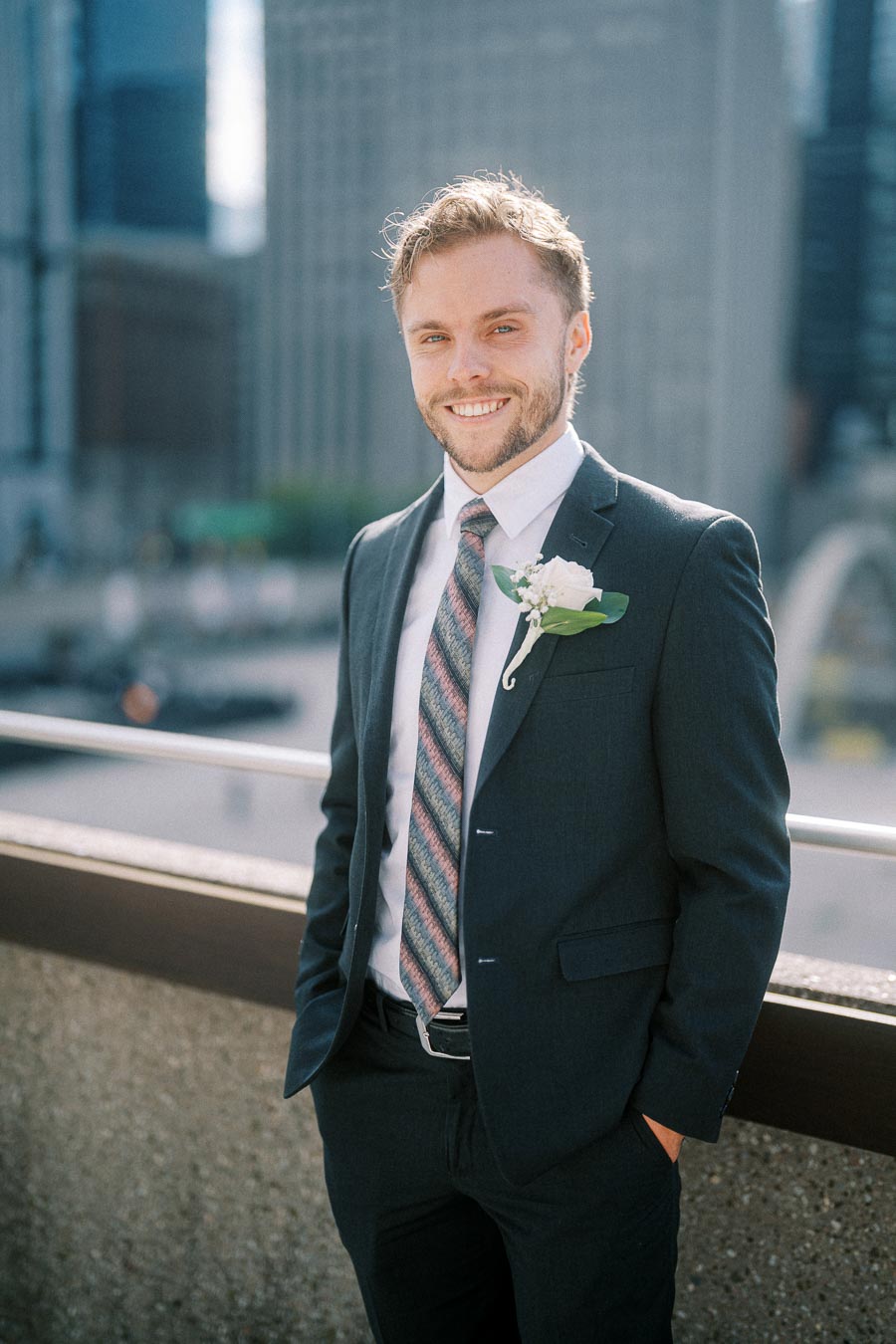 Smiling man in a dark suit with a patterned tie and boutonniere, standing on a city rooftop with blurred skyscrapers in the background.