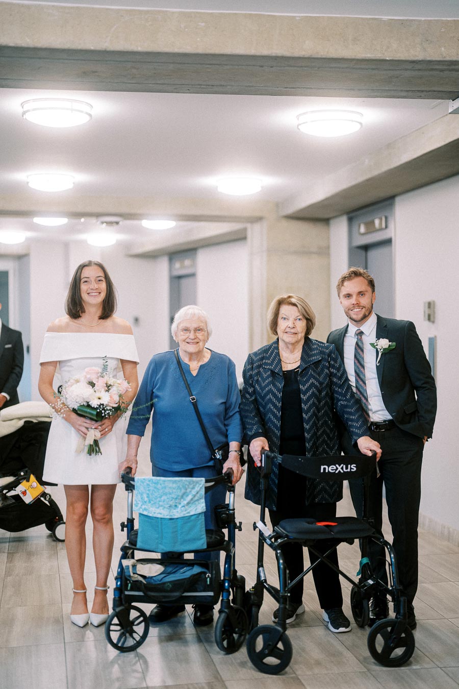 A young woman in a white dress holding a bouquet of flowers stands next to two elderly women using walkers and a man in a suit, all smiling indoors under bright lighting.