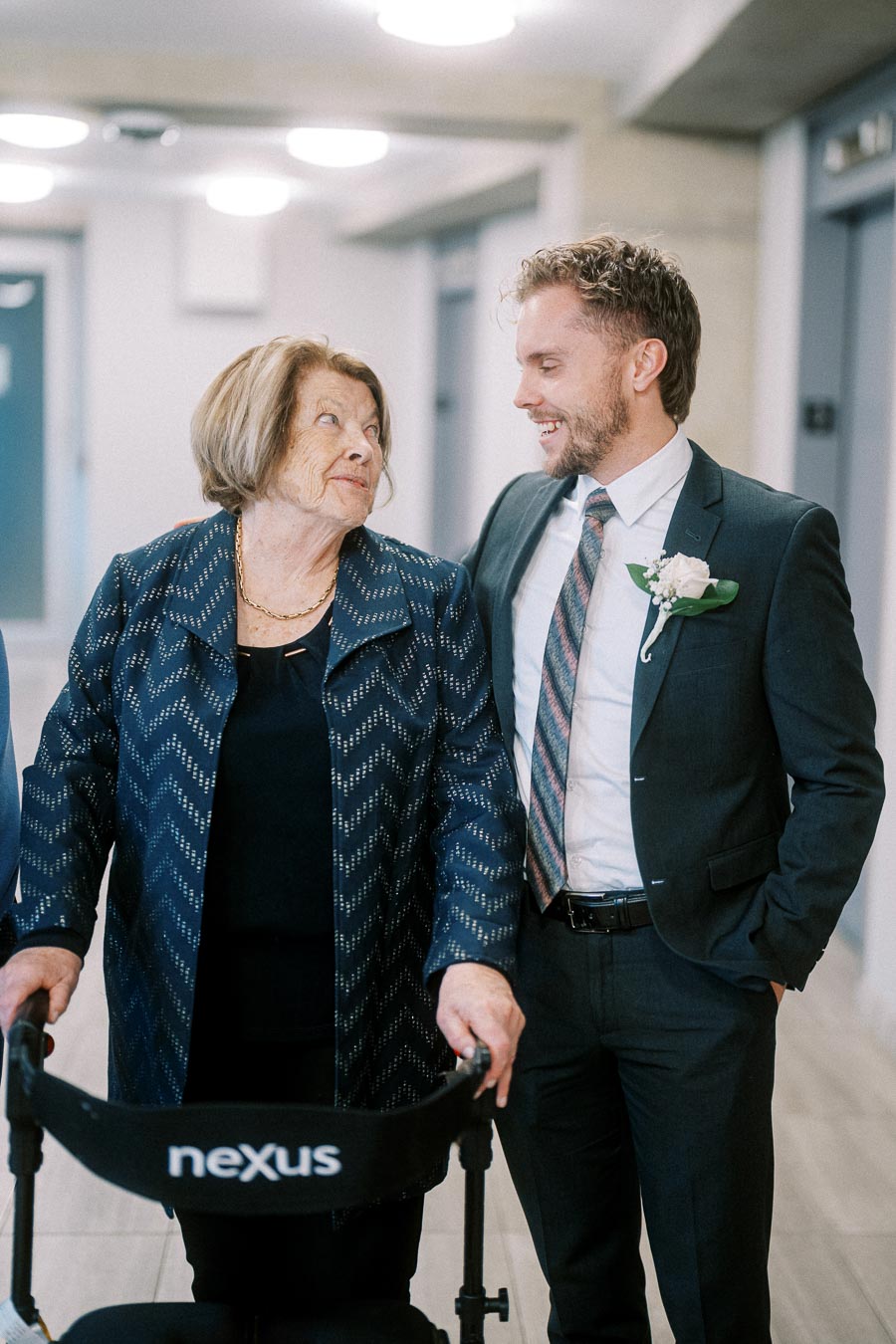 Elderly woman with a walker smiling at a man in a suit, standing in a hallway.