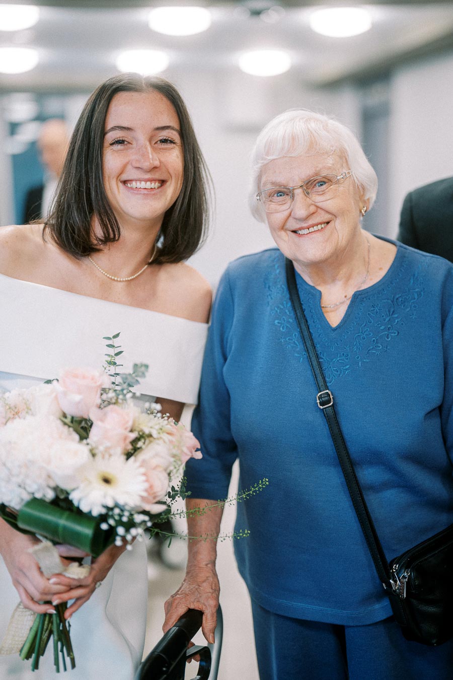 Smiling bride holding a bouquet stands beside an elderly woman in blue at a wedding event.