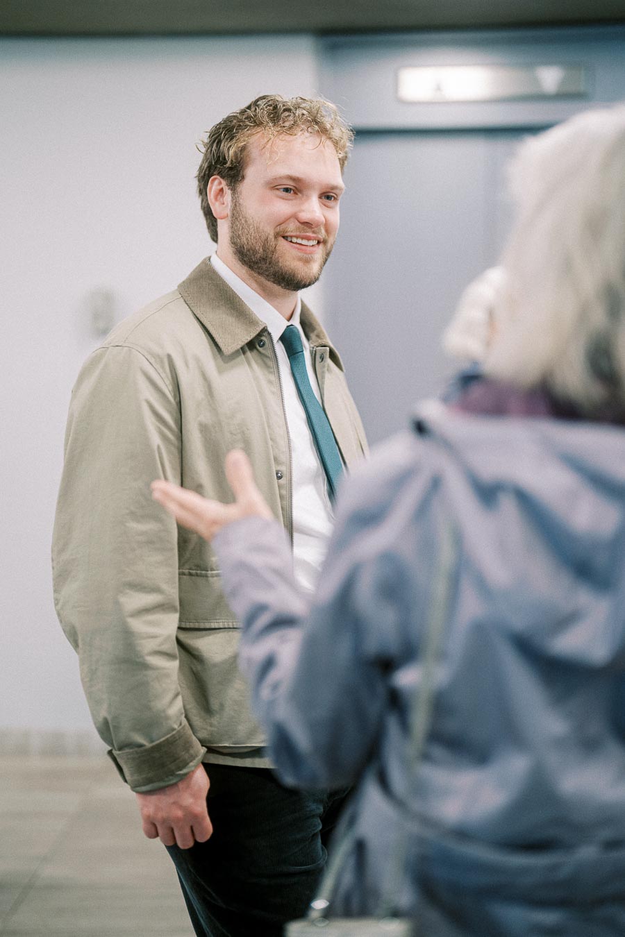 Man in beige jacket smiling while talking to a person in a blue coat inside a building.