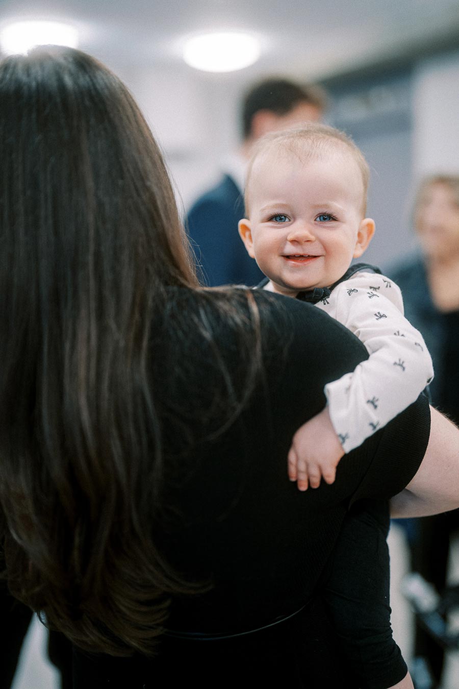 A baby smiling over the shoulder of a woman with long hair, wearing a patterned long-sleeve onesie, in an indoor setting with blurred people in the background.