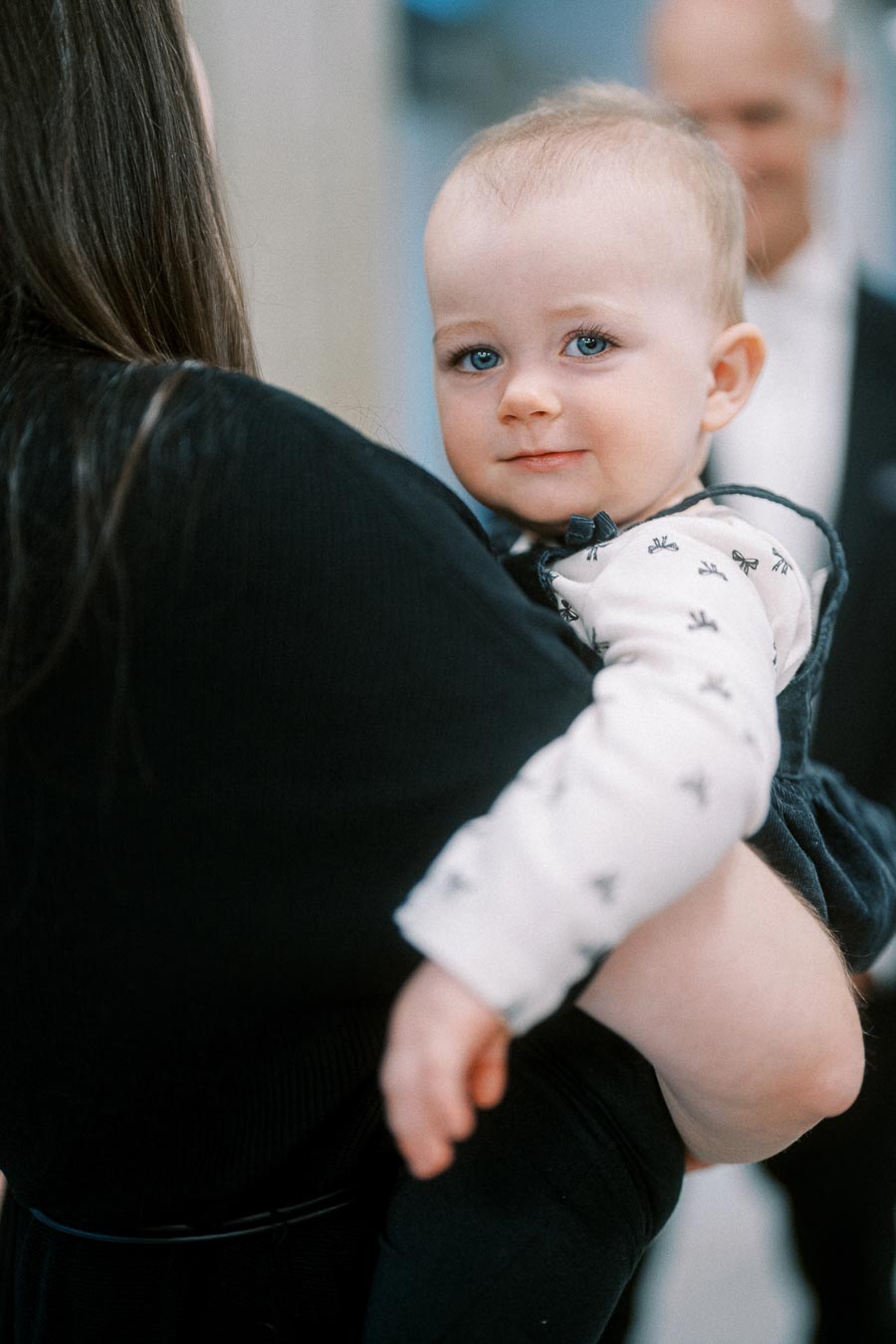 A smiling baby with blue eyes held by a person with long dark hair, wearing a patterned shirt and dark overalls, looking directly at the camera.
